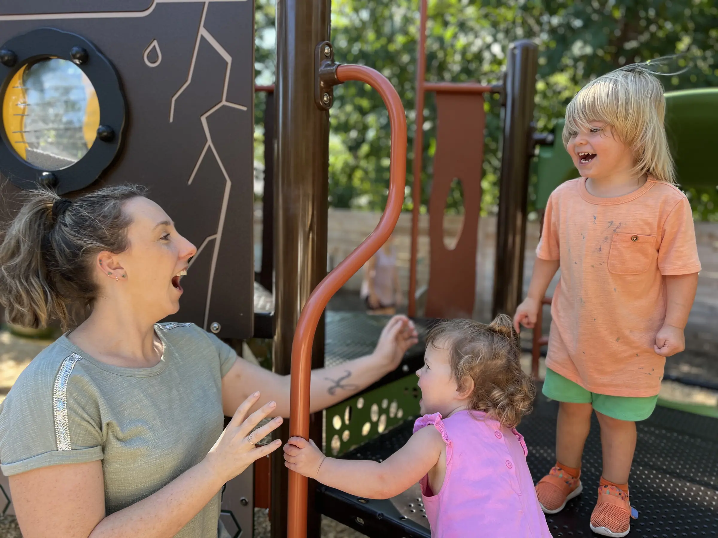 A Mockingbird teacher talking with two children on the shaded playscape