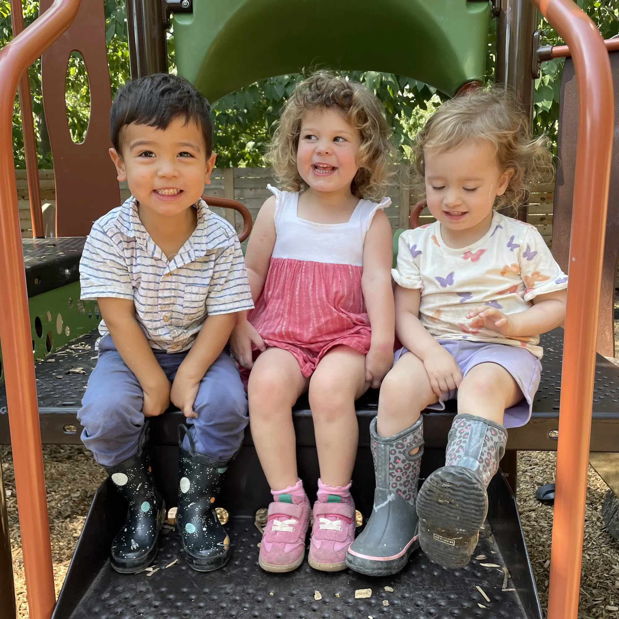 Three Mockingbird toddlers sharing a joyful moment on the shaded playscape